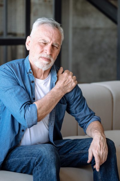 Senior man suffering from pain in shoulder Arthritis disease, problem with health. Senior man suffering from pain in shoulder, sitting on couch at home. Male touching, massaging inflammation zone.